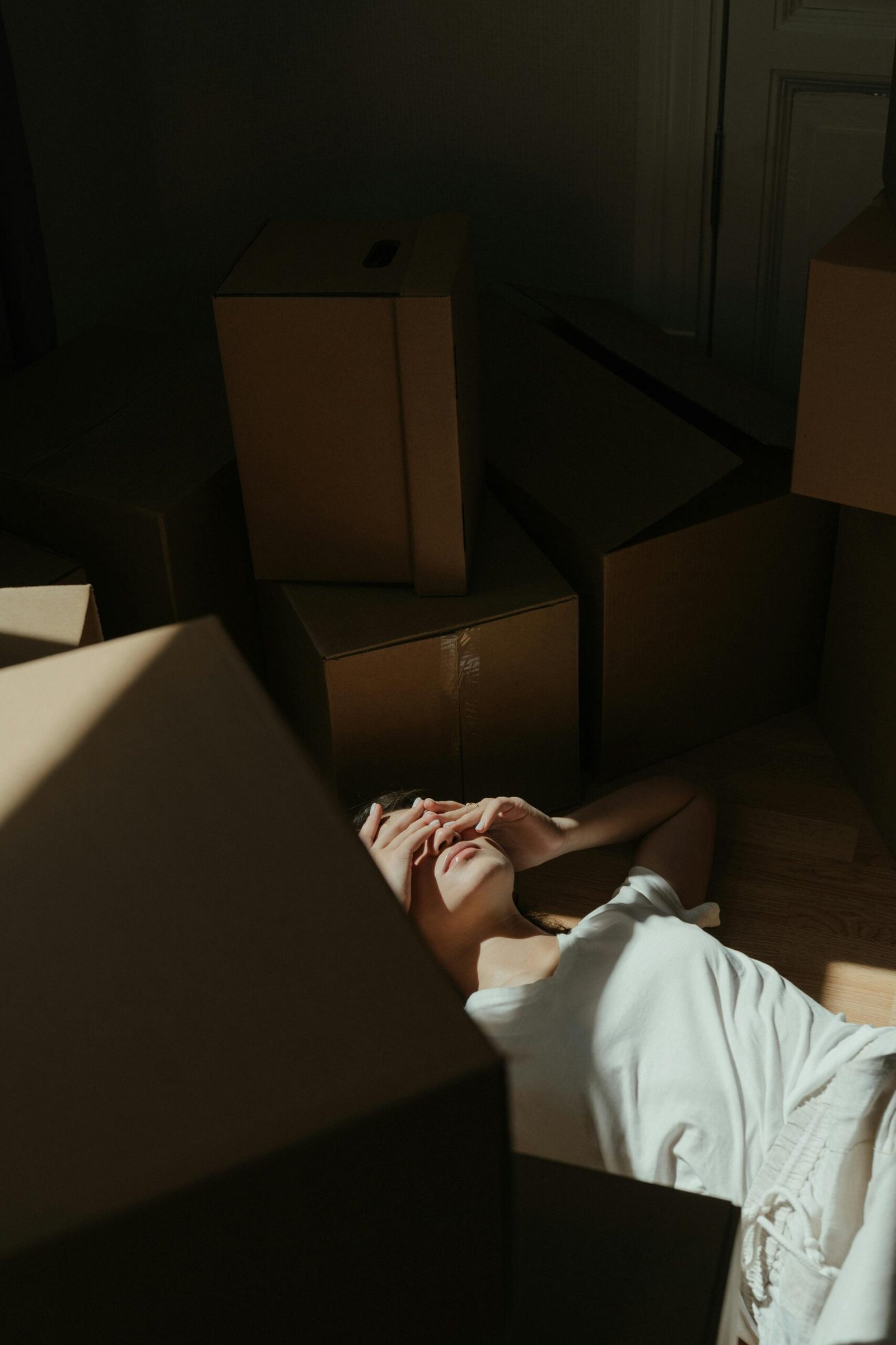 A person lying on the floor surrounded by cardboard boxes, shielding their face from sunlight, representing exhaustion and overwhelm—tomorrow is today.